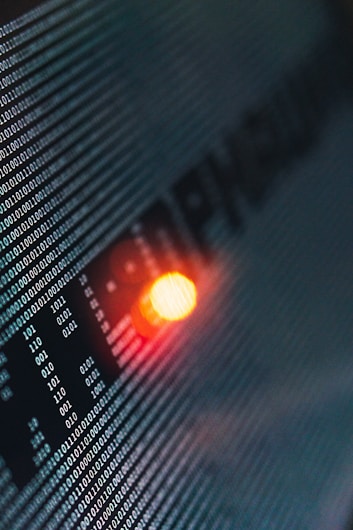 Binary code runs across a digital screen, highlighted by a glowing red-orange LED light. The numbers are aligned in a matrix pattern, creating a sense of digital complexity and data flow.