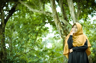 A person wearing a mustard-colored hijab and a black outfit is standing in a lush, green forest setting. They have a gentle smile and a contemplative pose, with one hand resting on their face. The background is filled with dense greenery and large tree trunks, creating a serene and natural atmosphere.