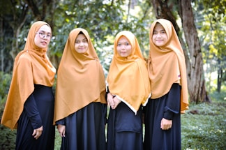 Four women wearing golden brown hijabs and long black dresses stand outdoors against a backdrop of green trees and grass.
