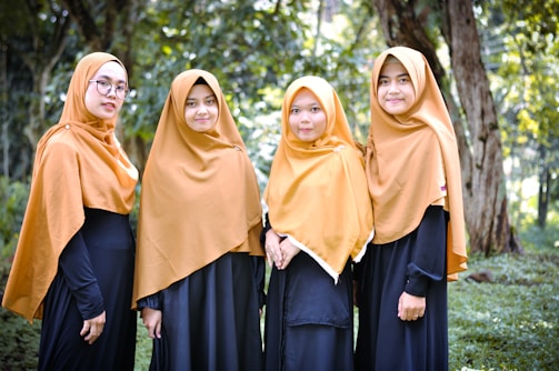 Four women wearing golden brown hijabs and long black dresses stand outdoors against a backdrop of green trees and grass.