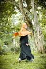 A person wearing a bright orange hijab and a long black dress is joyfully walking in a lush green forest. The surroundings include tall trees with textured bark and a carpet of green grass, creating a fresh and natural atmosphere.