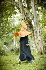 A person wearing a bright orange hijab and a long black dress is joyfully walking in a lush green forest. The surroundings include tall trees with textured bark and a carpet of green grass, creating a fresh and natural atmosphere.