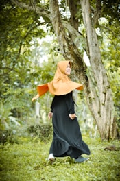 A person wearing a bright orange hijab and a long black dress is joyfully walking in a lush green forest. The surroundings include tall trees with textured bark and a carpet of green grass, creating a fresh and natural atmosphere.