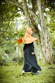 A person wearing a bright orange hijab and a long black dress is joyfully walking in a lush green forest. The surroundings include tall trees with textured bark and a carpet of green grass, creating a fresh and natural atmosphere.