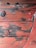 Weathered wooden surface with peeling red paint, revealing aged wood beneath. Large round metal bolts secure the planks.