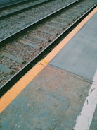 A section of a railway track bordered by a platform with a visible yellow safety line separating the two. The track is covered with gravel and wooden sleepers supporting the metal rails, showing signs of use and weather exposure. The platform surface appears worn with some discoloration and cracks.