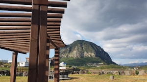 A wooden pergola is in the foreground with a picturesque view of a large, lush green mountain in the background. The sky is partly cloudy, adding a dramatic effect to the scene. There are a few small buildings and open fields visible near the base of the mountain.