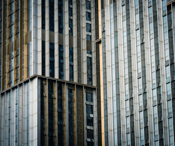 Tall buildings with modern architectural design feature a pattern of vertical and horizontal lines, composed of glass and metal panels. The facade displays a gradient of colors from light to dark, highlighting the structural elements and windows.
