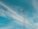 A tall metal communication tower stands against a bright blue sky with wispy white clouds. The structure includes antennas and dishes at the top, used for wireless communication.