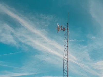 A tall metal communication tower stands against a bright blue sky with wispy white clouds. The structure includes antennas and dishes at the top, used for wireless communication.