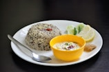 A plate containing a serving of rice with lentils, garnished with spices and herbs, accompanied by a yellow bowl of yogurt-based dip with visible spices. Slices of lime and cucumber along with a green chili and a small portion of chutney are placed on the plate. A spoon rests beside the rice on the white plate, all set against a dark background.