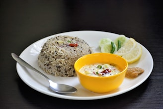 A plate containing a serving of rice with lentils, garnished with spices and herbs, accompanied by a yellow bowl of yogurt-based dip with visible spices. Slices of lime and cucumber along with a green chili and a small portion of chutney are placed on the plate. A spoon rests beside the rice on the white plate, all set against a dark background.