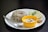 A plate containing a serving of rice with lentils, garnished with spices and herbs, accompanied by a yellow bowl of yogurt-based dip with visible spices. Slices of lime and cucumber along with a green chili and a small portion of chutney are placed on the plate. A spoon rests beside the rice on the white plate, all set against a dark background.