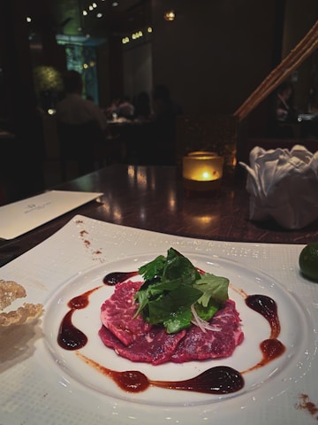 A gourmet dish is elegantly presented on a white plate. The centerpiece is thin slices of marbled red meat, garnished with fresh green herbs. The meat is surrounded by artistic drizzles of a dark brown sauce. The table setting includes a lit candle, adding a warm ambiance to the dimly lit restaurant setting with other patrons visible in the background.