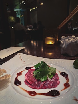 A gourmet dish is elegantly presented on a white plate. The centerpiece is thin slices of marbled red meat, garnished with fresh green herbs. The meat is surrounded by artistic drizzles of a dark brown sauce. The table setting includes a lit candle, adding a warm ambiance to the dimly lit restaurant setting with other patrons visible in the background.