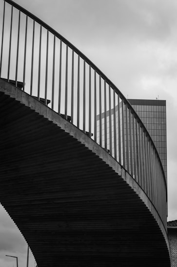 A striking black and white photograph featuring the curved form of a modern bridge with vertical railings set against an overcast sky. In the background, a tall glass building with a grid pattern is partially visible, adding an urban element to the composition.