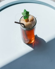 A tall glass filled with an iced tea-like beverage topped with frothy bubbles, adorned with fresh mint leaves and two lychee fruits on a skewer. A straw is placed in the drink, and the glass is set on a white surface with a soft shadow cast across it.