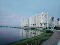 A large residential complex with several multi-story buildings is situated along the edge of a calm body of water. Tall palm trees line the walkway near the buildings. The water reflects the structures, creating a serene and mirrored effect. The sky is mostly overcast with subtle hints of pink, indicating a possible sunrise or sunset. The foreground shows a paved path and some greenery lining the water's edge.