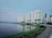 A large residential complex with several multi-story buildings is situated along the edge of a calm body of water. Tall palm trees line the walkway near the buildings. The water reflects the structures, creating a serene and mirrored effect. The sky is mostly overcast with subtle hints of pink, indicating a possible sunrise or sunset. The foreground shows a paved path and some greenery lining the water's edge.