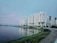 A large residential complex with several multi-story buildings is situated along the edge of a calm body of water. Tall palm trees line the walkway near the buildings. The water reflects the structures, creating a serene and mirrored effect. The sky is mostly overcast with subtle hints of pink, indicating a possible sunrise or sunset. The foreground shows a paved path and some greenery lining the water's edge.