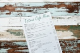 A rustic menu with the header 'Tsitrus Café Menu,' featuring various food and drink options such as pizzas, savory pancakes, pot pies, cheesecakes, pancakes, ice cream, cold drinks, and hot drinks. The menu is placed at an angle on a distressed wooden surface with peeling paint in shades of white, teal, and brown.