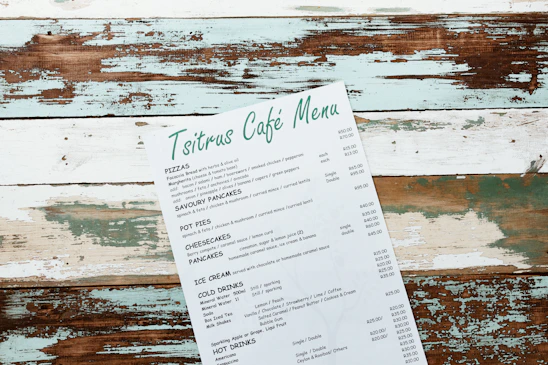 A rustic menu with the header 'Tsitrus Café Menu,' featuring various food and drink options such as pizzas, savory pancakes, pot pies, cheesecakes, pancakes, ice cream, cold drinks, and hot drinks. The menu is placed at an angle on a distressed wooden surface with peeling paint in shades of white, teal, and brown.