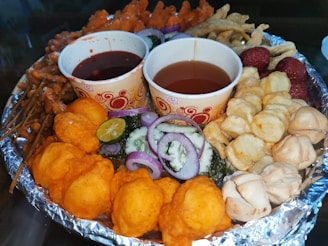 A platter filled with various fried snacks, including orange puffed items, skewered meats, and crispy chips. Two paper cups contain dipping sauces. Sliced onions, cucumbers, and a lime wedge garnish the dish for added freshness.