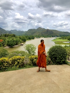 A monk dressed in orange robes walks along a path with a serene river flowing behind him. The background features lush greenery and distant mountains under a cloudy sky, giving a tranquil and natural setting.