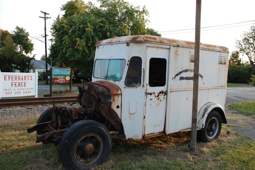 An old, rusty delivery truck is parked beside railroad tracks. The vehicle is missing its front engine cover, revealing mechanical components. The white paint is peeling and rusted, with a faded design on the side. In the background, there are trees, a fence, and a sign advertising a fence company.