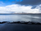 A serene seascape with calm waters in the foreground, bordered by dark rocks. In the distance, a snowy mountain range is partially obscured by a cloud-filled sky, creating a dramatic contrast between the bright snow and the grey clouds.