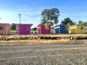 Four small, vividly colored houses line up under a bright blue sky. Each house is painted a different color: orange, pink, blue, and yellow. They are set against a backdrop of lush greenery with trees visible behind the homes. In front of the houses, there is a dirt road with scattered patches of grass and some mud puddles.