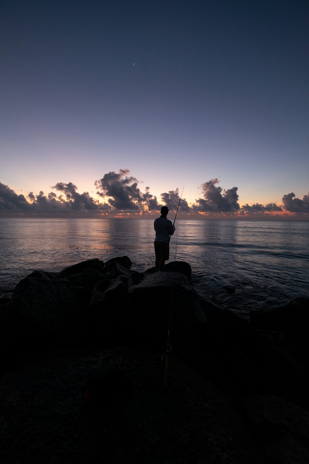 silhouette of man standing on rock near body of water during sunset