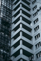 Tall modern building with geometric architectural design, featuring prominent balconies and a glass facade on one side. The atmosphere appears urban and slightly austere.