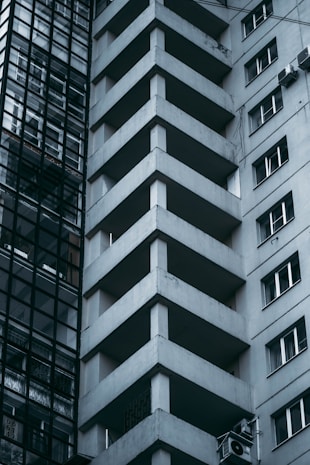 Tall modern building with geometric architectural design, featuring prominent balconies and a glass facade on one side. The atmosphere appears urban and slightly austere.