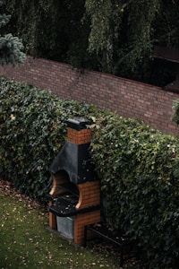 A brick barbecue situated in a garden setting with dense foliage surrounding it. The barbecue has a distinct chimney and a small bench is placed beside it. The ground is covered with a well-maintained lawn and a few fallen leaves.