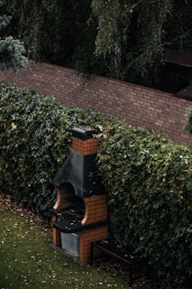 A brick barbecue situated in a garden setting with dense foliage surrounding it. The barbecue has a distinct chimney and a small bench is placed beside it. The ground is covered with a well-maintained lawn and a few fallen leaves.