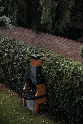 A brick barbecue situated in a garden setting with dense foliage surrounding it. The barbecue has a distinct chimney and a small bench is placed beside it. The ground is covered with a well-maintained lawn and a few fallen leaves.