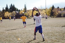 A group of young soccer players is on a grassy field. One player in a white jersey with '11' printed on the back is preparing to throw the ball. Other players are wearing yellow jerseys. The backdrop consists of trees with autumn foliage and a residential area.