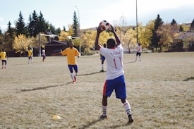 A group of young soccer players is on a grassy field. One player in a white jersey with '11' printed on the back is preparing to throw the ball. Other players are wearing yellow jerseys. The backdrop consists of trees with autumn foliage and a residential area.