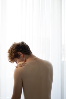 A person with light skin and curly hair is shown from the back, pressing their left hand on their right shoulder. The background is softly illuminated by sheer white curtains, creating a gentle, diffused light.
