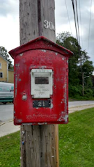 A vintage red fire alarm box affixed to a wooden utility pole, displaying signs of weathering and peeling paint. Behind it is a residential neighborhood featuring a house and a van, along with a lush green lawn and road.