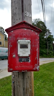 A vintage red fire alarm box affixed to a wooden utility pole, displaying signs of weathering and peeling paint. Behind it is a residential neighborhood featuring a house and a van, along with a lush green lawn and road.