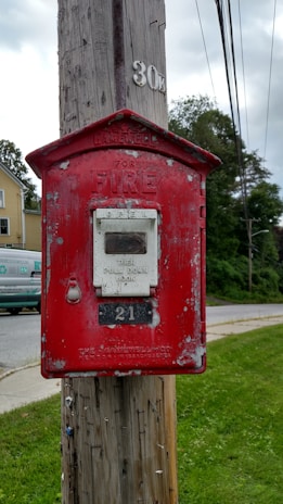 A vintage red fire alarm box affixed to a wooden utility pole, displaying signs of weathering and peeling paint. Behind it is a residential neighborhood featuring a house and a van, along with a lush green lawn and road.