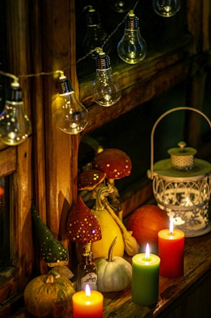 A cozy and warm scene featuring lit candles in orange, green, and red colors, surrounded by decorative gourds and a small white pumpkin. There is a whimsical touch with toadstool figurines adorned with glitter, a small glass bottle, and a decorative lantern adding to the warm ambiance. The setting is further illuminated by a string of large vintage-style bulbs, creating a soft glow against a wooden window frame.