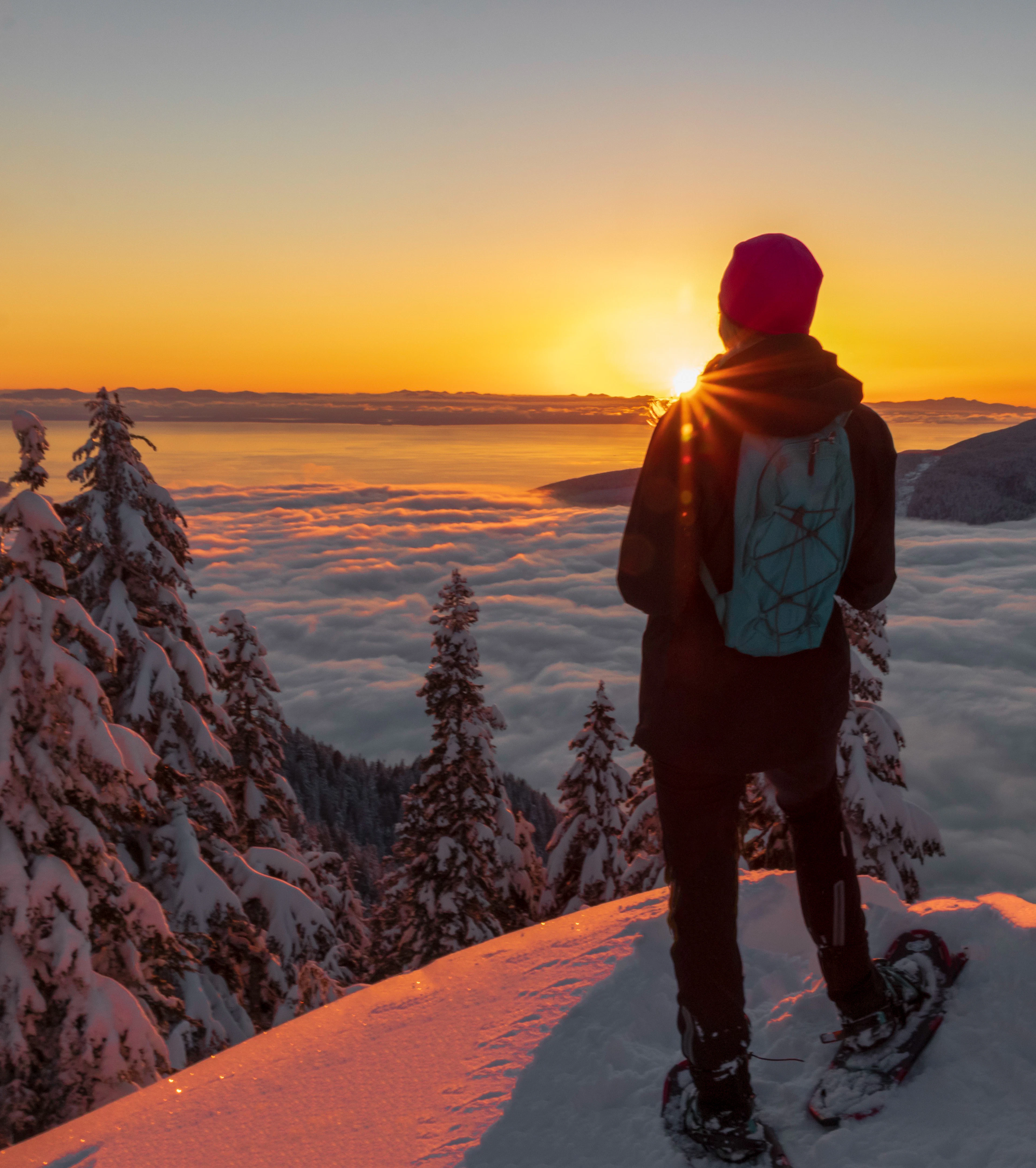 A snowshoer at a mountain summit near Vancouver watches the sun set over a sea of clouds.