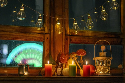 A cozy indoor scene with a wooden window adorned with string lights featuring exposed bulbs. On the windowsill, there are four lit candles in red, green, and yellow colors. A decorative lantern, mushroom ornaments, a small bottle, and a metal cup are also present. The window shows a blurred colorful light display outside with a glowing rainbow-like pattern.