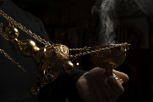 A hand holding an ornate, golden incense burner that is emitting smoke, set against a dark background.