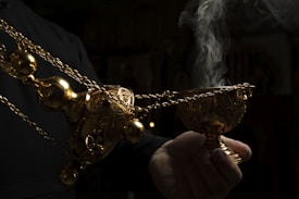 A hand holding an ornate, golden incense burner that is emitting smoke, set against a dark background.
