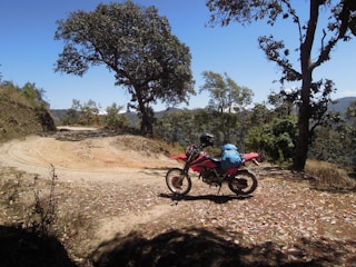A red motorcycle is parked on a dirt path surrounded by trees. The path is lined with fallen leaves, and the background features a clear blue sky and distant hills. The motorcycle has a blue backpack strapped to it and a helmet resting on the seat.