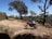 A red motorcycle is parked on a dirt path surrounded by trees. The path is lined with fallen leaves, and the background features a clear blue sky and distant hills. The motorcycle has a blue backpack strapped to it and a helmet resting on the seat.